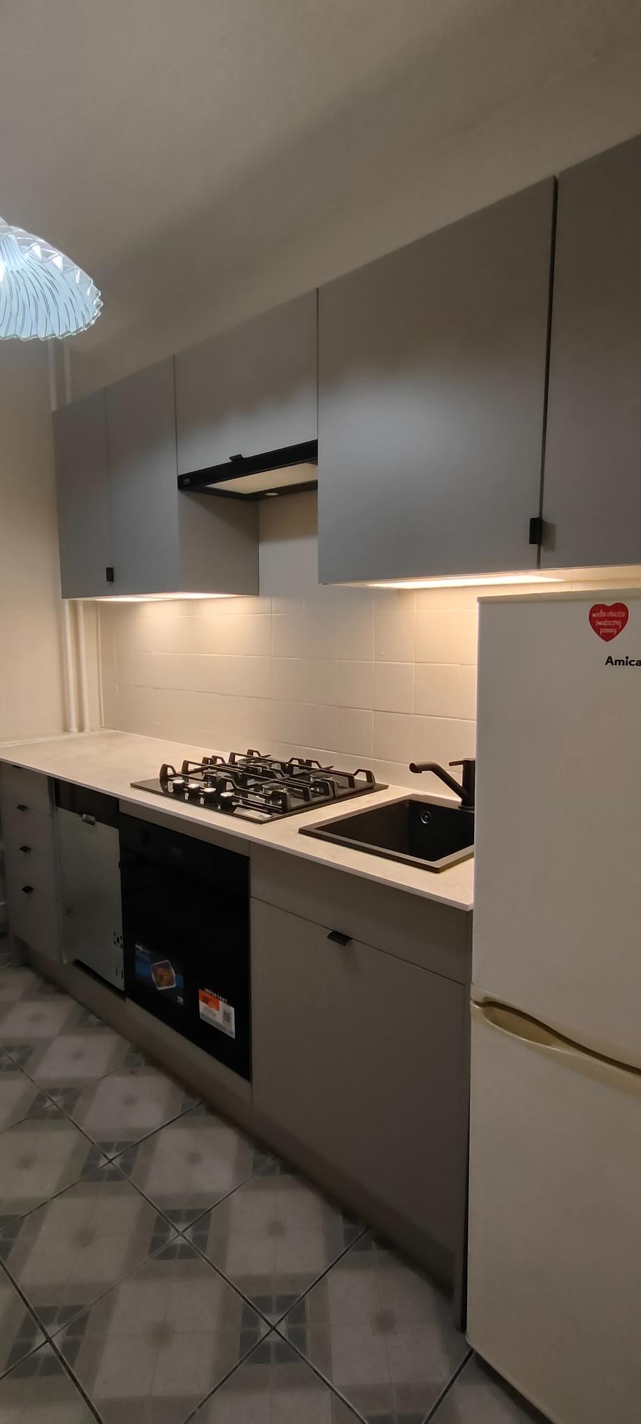 Szara kuchnia po remoncie z czarnym zlewem i płytą gazową. Widoczne szafki wiszące i stojące, lodówka Amica. Ukończony remont kuchni z nowymi meblami i sprzętami.