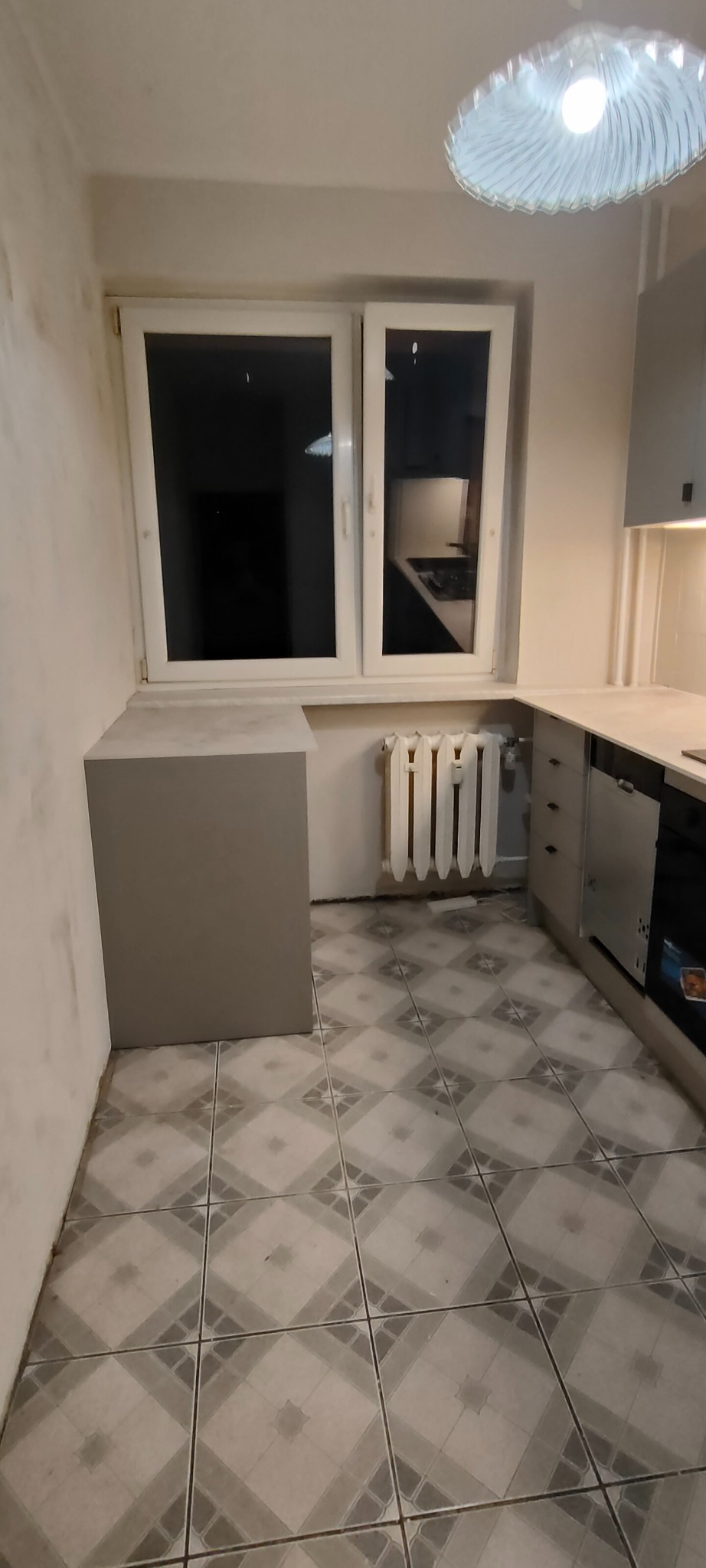 Fragment kuchni w trakcie remontu: szare szafki, okno, grzejnik i wzorzysta podłoga. Ściany przygotowane do malowania. Widoczny etap prac wykończeniowych.