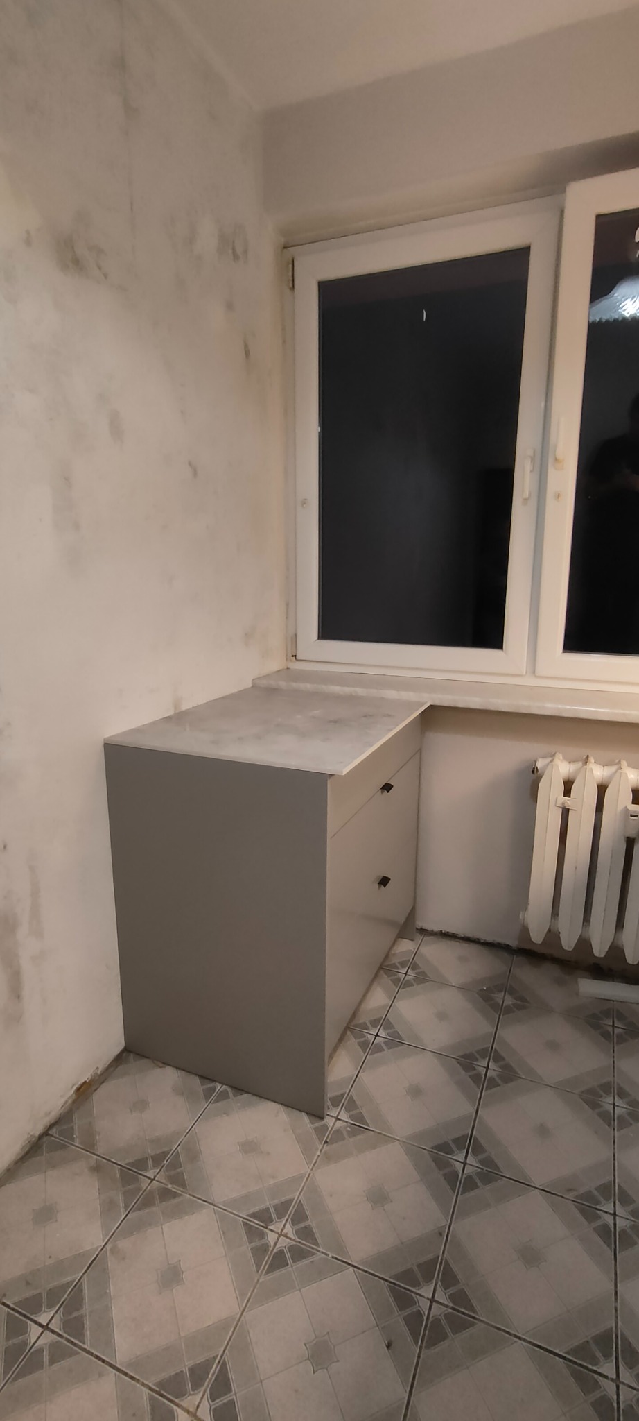 Szara szafka z szufladami i blatem, dopasowana do wnęki okiennej. Widoczny grzejnik i płytki podłogowe. Ściana z widocznymi śladami wilgoci.