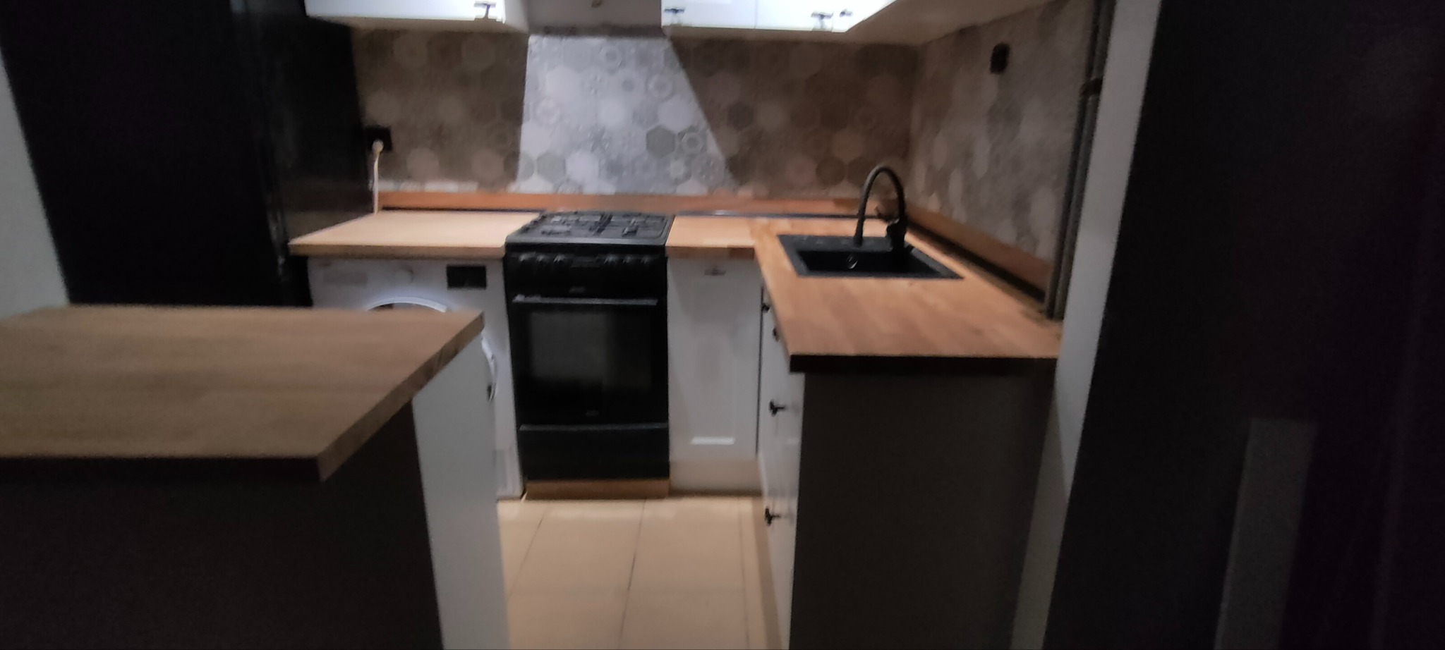 Nowoczesna kuchnia po remoncie: białe szafki z drewnianymi blatami, czarny zlew i kuchenka. Heksagonalne płytki na ścianie. Pralka w zabudowie.