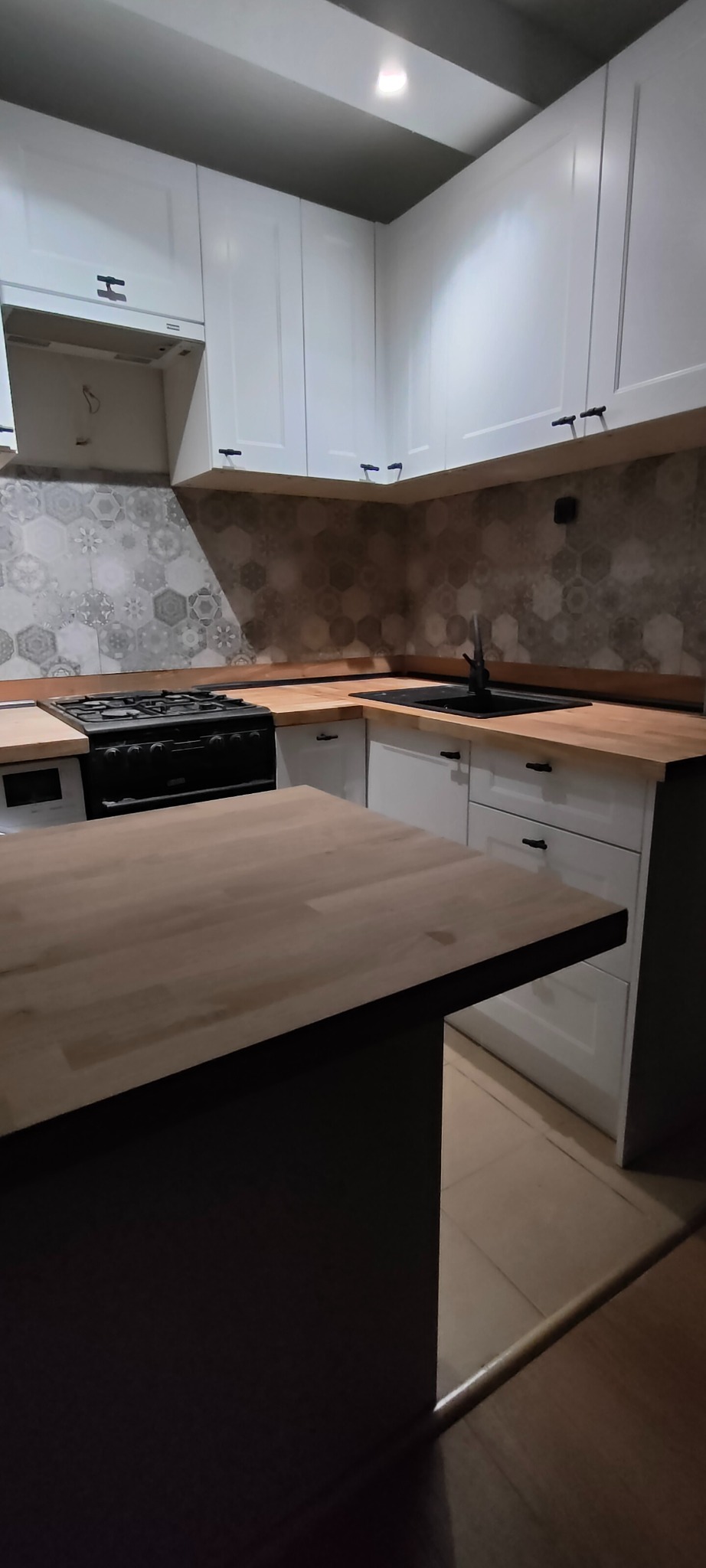 Nowoczesna kuchnia z białymi szafkami, drewnianym blatem i heksagonalnymi płytkami na ścianie. Czarny zlew i kuchenka dodają kontrastu. Widoczna wyspa kuchenna.