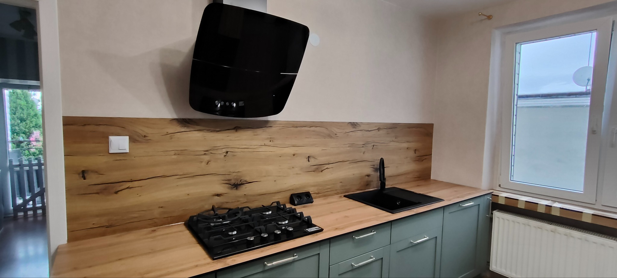 Nowoczesna kuchnia z drewnianym panelem ściennym, czarnym okapem i płytą gazową. Uwagę przykuwają zielone szafki i czarna armatura, tworzące spójną całość.