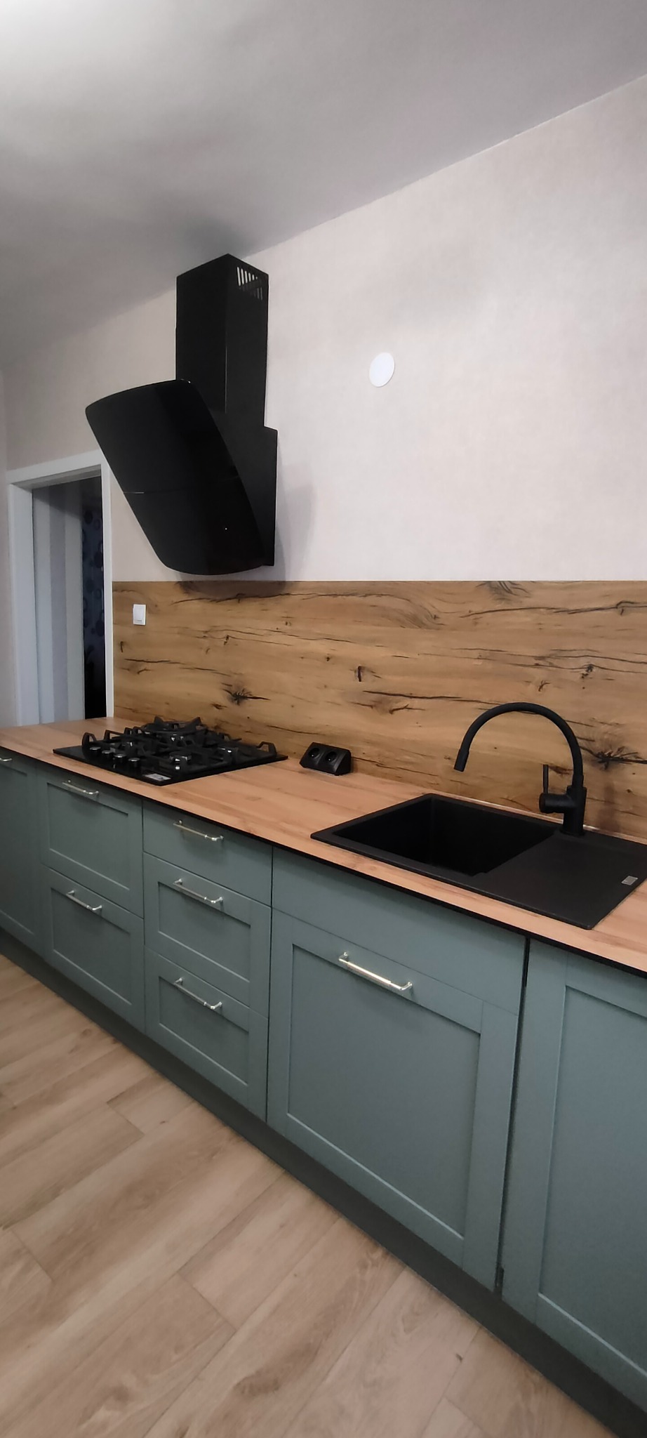 Nowoczesna kuchnia z szarymi szafkami, drewnianym blatem i czarnym zlewem oraz płytą gazową. Uwagę przykuwa czarny okap nad płytą.