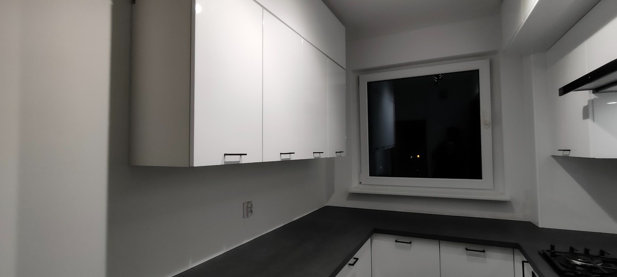 Biała, nowoczesna kuchnia z górnymi szafkami na wysoki połysk i czarnymi uchwytami. Ciemny blat i okno z widokiem na nocne miasto. Minimalistyczny design.