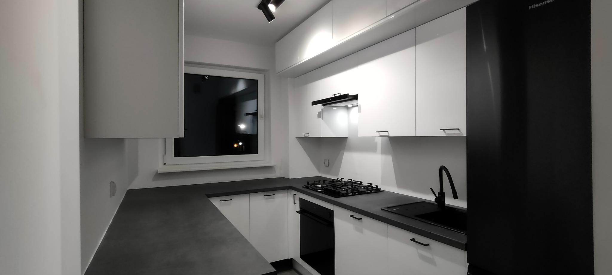 Nowoczesna kuchnia w bieli i czerni. Minimalistyczne szafki, czarny blat i sprzęt AGD. Oświetlenie punktowe podkreśla detale. Widok na okno z ciemnym otoczeniem.