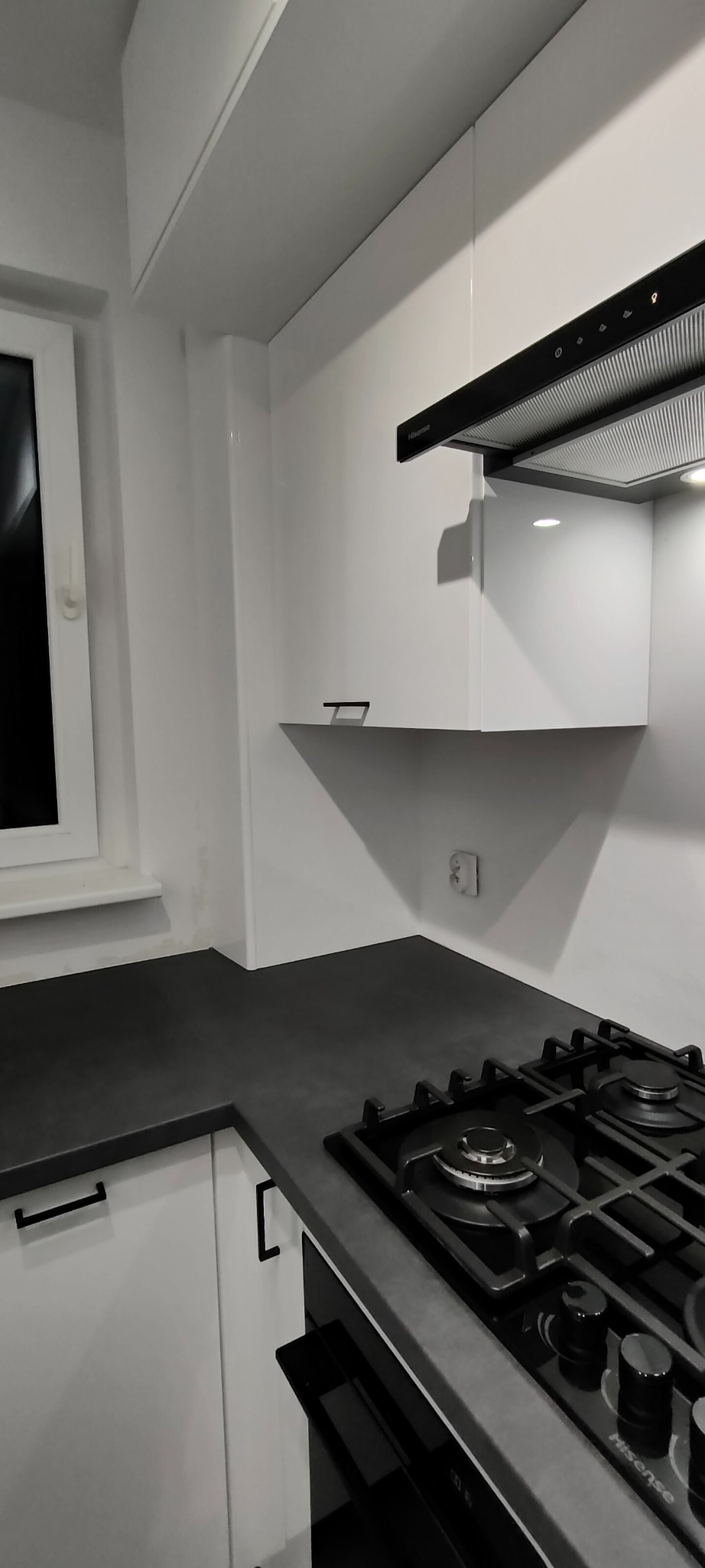 Nowoczesna kuchnia z białymi szafkami, czarnym blatem i płytą gazową. Minimalistyczny design, oświetlenie podszafkowe, gładkie fronty. Widok na fragment okna.