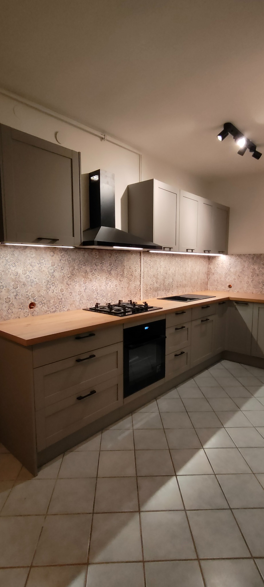 Szara kuchnia w kształcie litery L z drewnianym blatem, czarnym okapem i płytą gazową. Nad blatem dekoracyjne płytki. Oświetlenie podszafkowe LED.