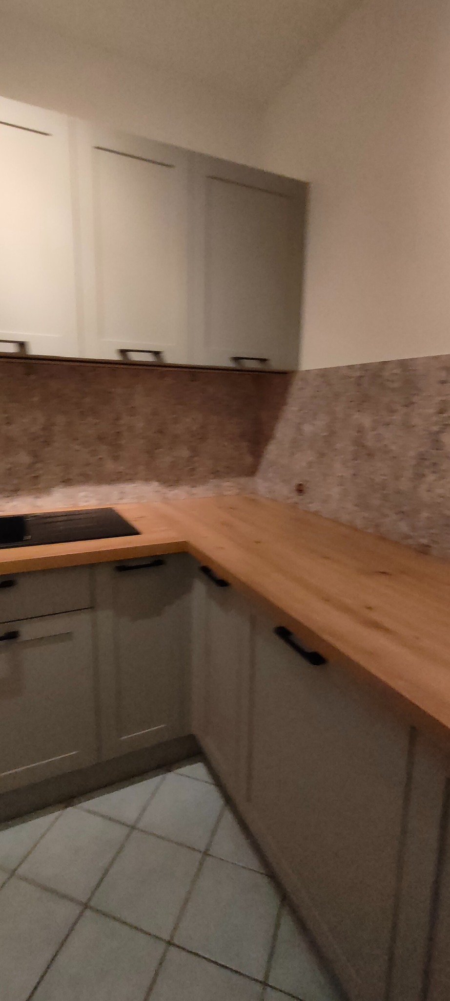 Jasna kuchnia z drewnianym blatem i szarymi szafkami. Ujęcie z niskiego kąta, widoczna płyta indukcyjna i fragment podłogi z płytek.