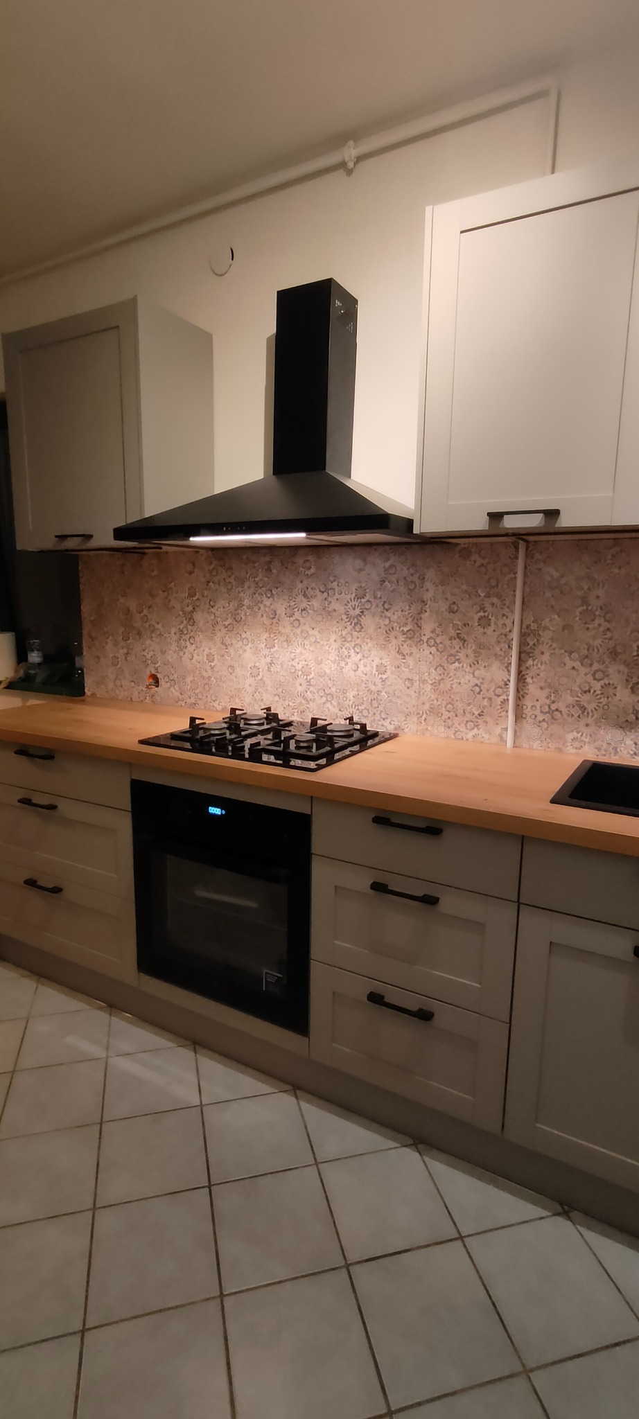 Jasna kuchnia z drewnianym blatem, czarnym okapem i płytą gazową. Fronty szafek w stylu klasycznym z czarnymi uchwytami. Płytki nad blatem w delikatny wzór.