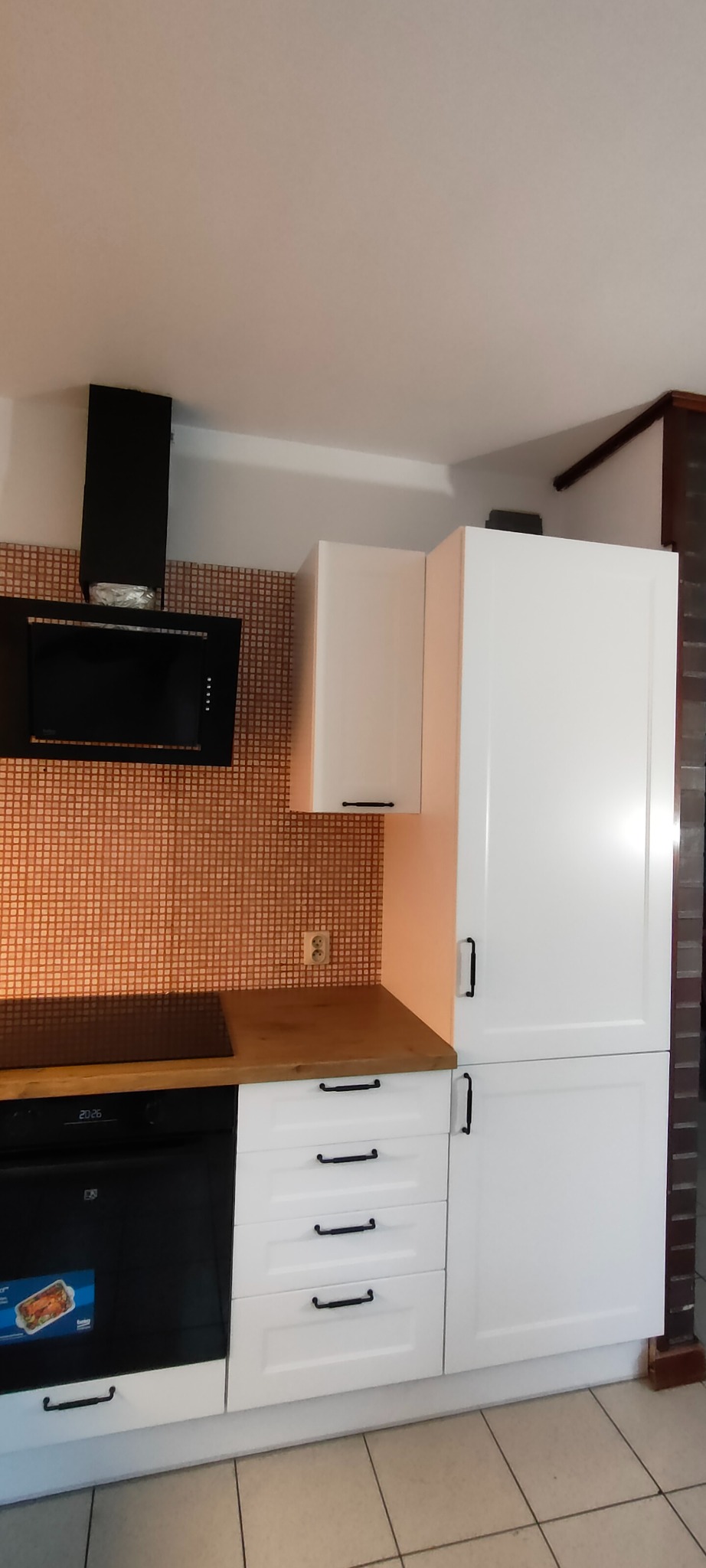 Biała zabudowa kuchenna z czarnymi uchwytami, brązowy blat i mozaika na ścianie. Widoczny piekarnik i okap. Ujęcie z dołu, prezentujące detale wykończenia.