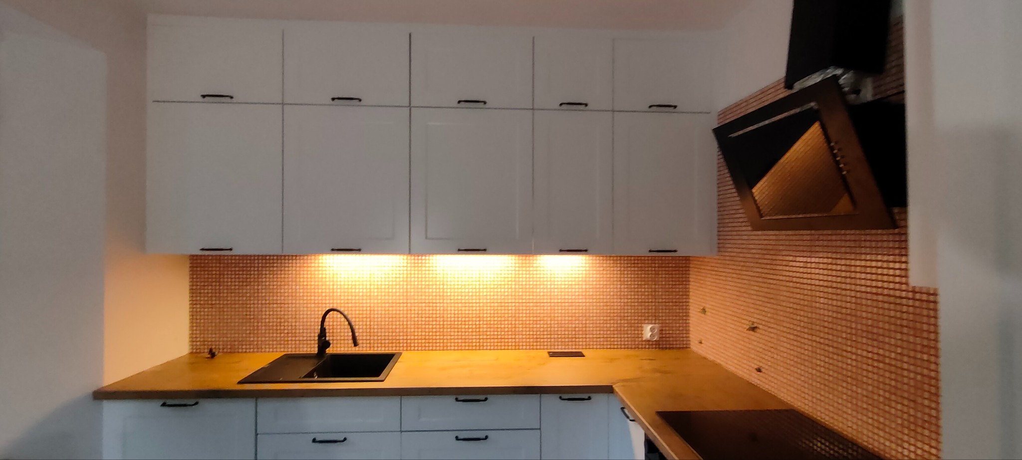 Nowoczesna kuchnia z białymi szafkami, drewnianym blatem i mozaiką na ścianie. Czarny zlew i okap. Oświetlenie podszafkowe dodaje ciepła. Minimalistyczny design.