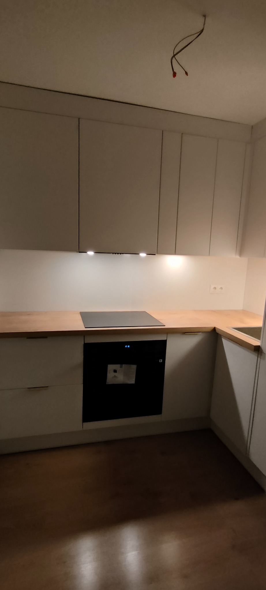Jasna kuchnia z drewnianym blatem i czarną płytą indukcyjną. Minimalistyczny design, górne szafki z oświetleniem punktowym. Widoczne niedokończone podłączenie elektryczne.