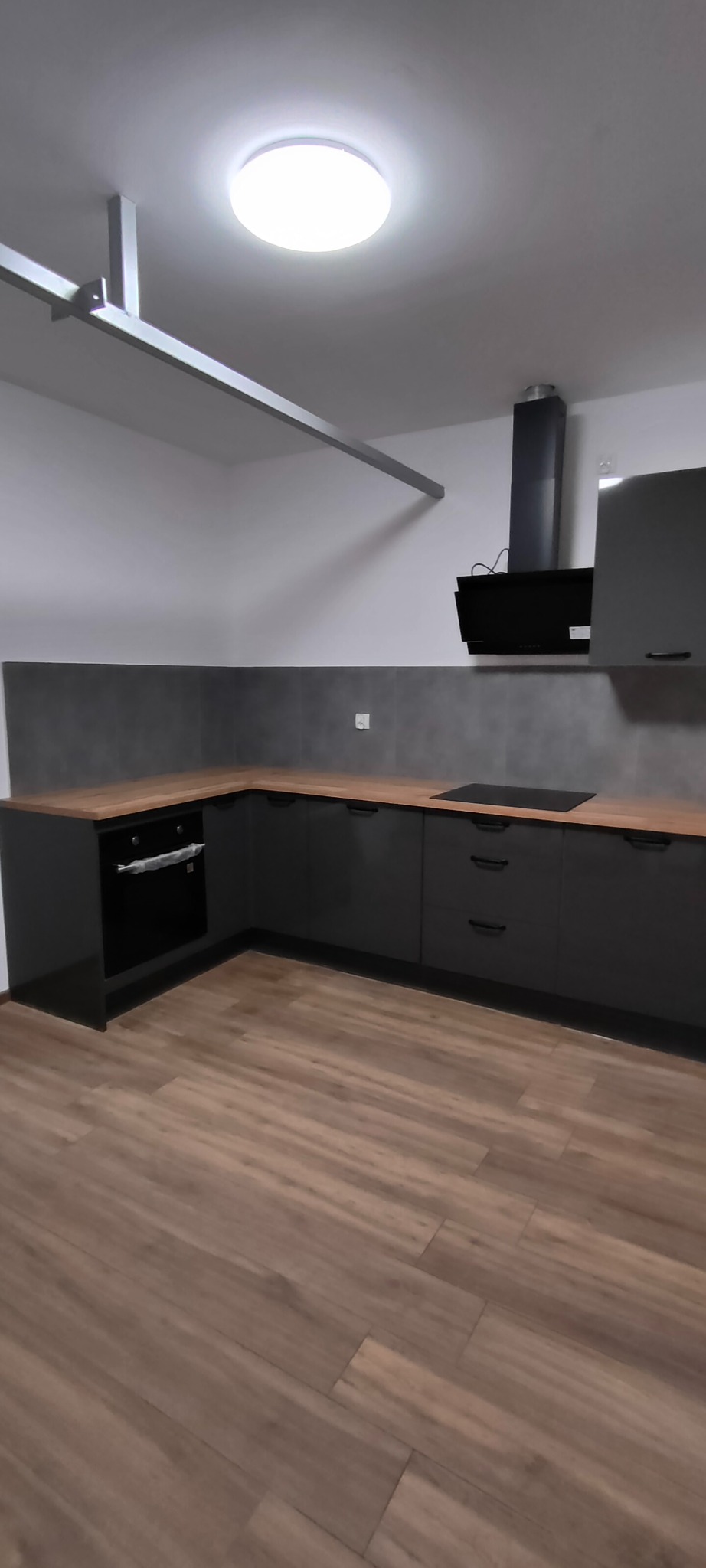 Nowoczesna, szara kuchnia z drewnianym blatem i czarnym okapem. Ujęcie z perspektywy podłogi, widoczne oświetlenie sufitowe i fragment instalacji.