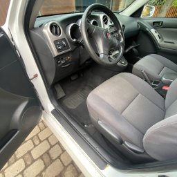 Czyścioch z dojazdem Mobilne sprzatanie - Wnętrze białego samochodu z otwartymi drzwiami kierowcy, widoczne szare fotele, deska rozdzielcza i fragment kostki brukowej na zewnątrz. Ujęcie z bliska, naturalne światło.