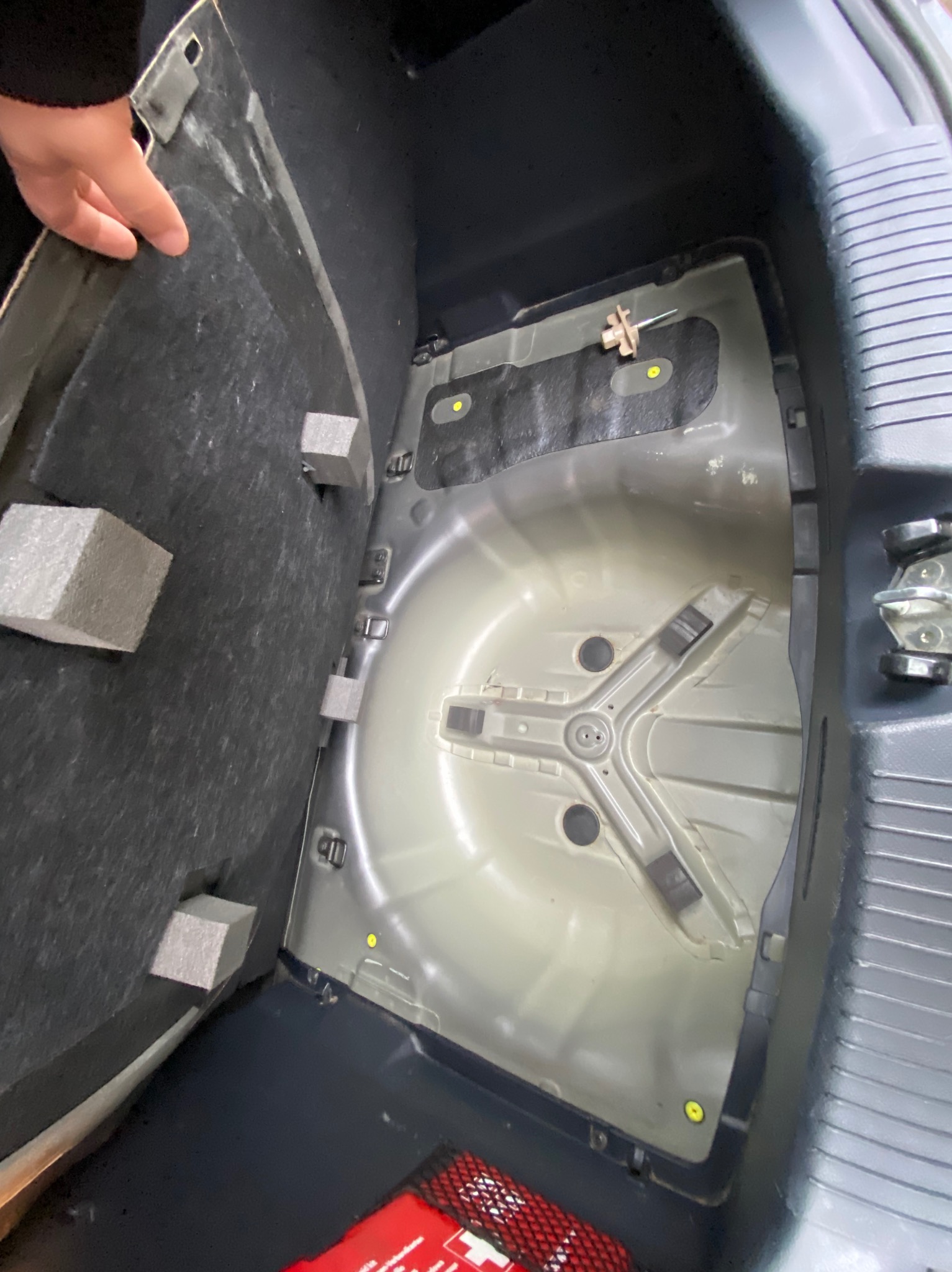 Otwarty bagażnik samochodowy z widocznym kołem zapasowym i elementami mocującymi. Widoczna ręka podnosząca element wykończenia. Czyste wnętrze bagażnika.