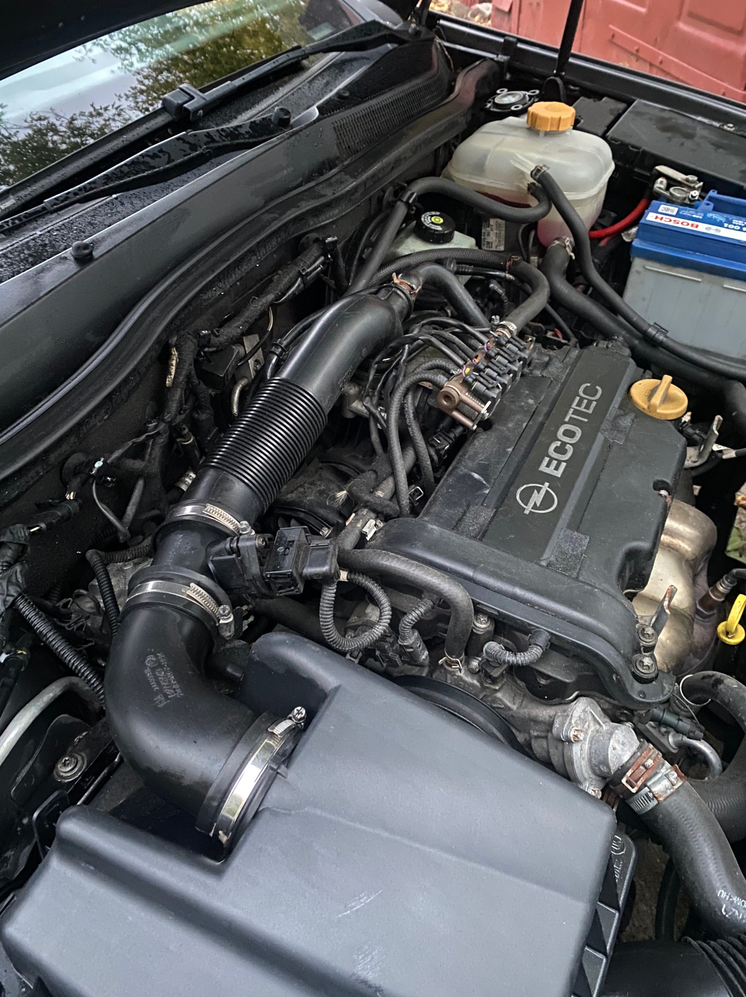 Silnik samochodu marki Opel, widoczne elementy takie jak akumulator, przewody, filtr powietrza. Silnik Ecotec.