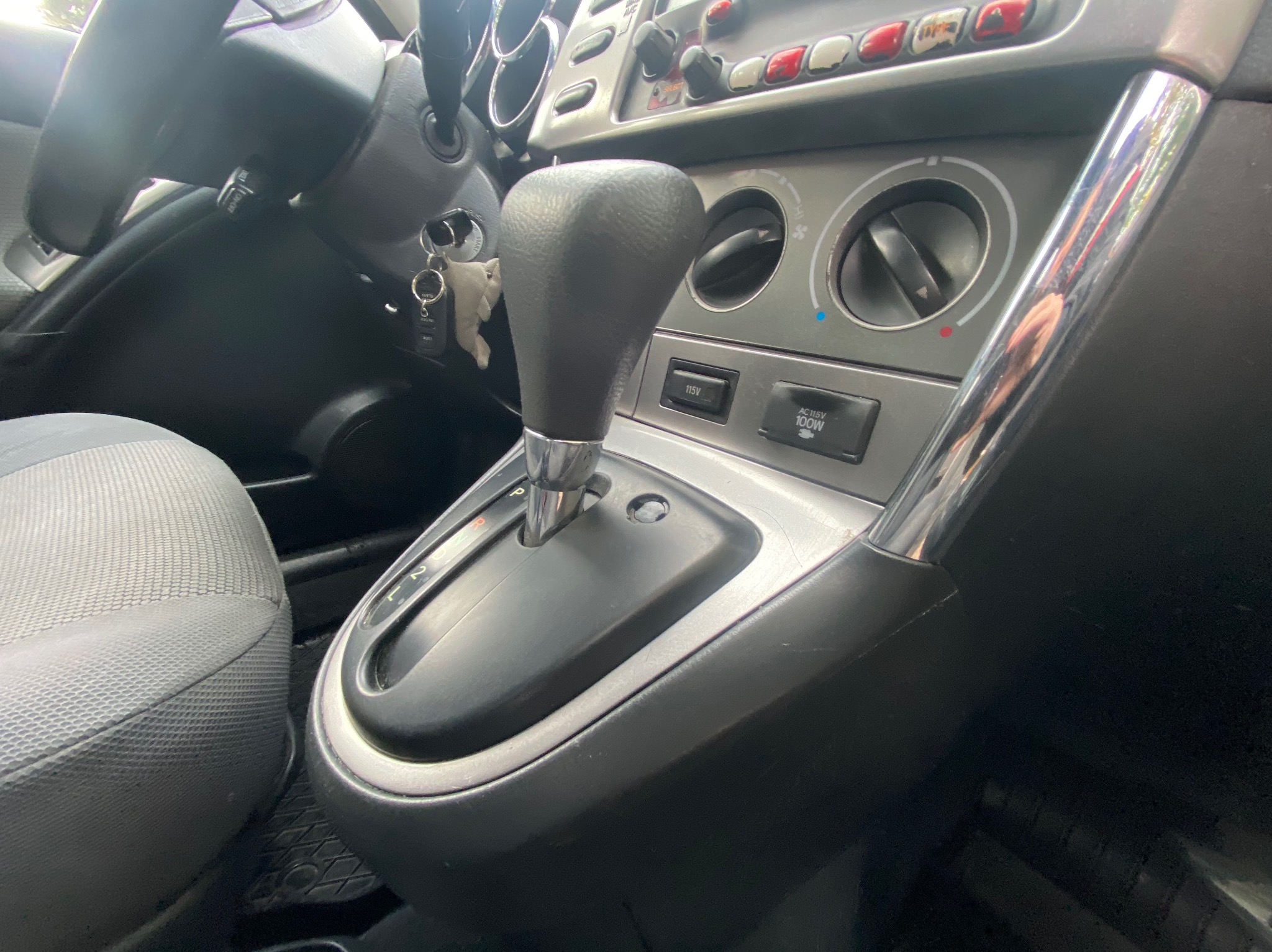 Wnętrze samochodu z dźwignią zmiany biegów, widoczne detale deski rozdzielczej, w tym pokrętła regulacji i gniazda zasilania. Kluczyki wiszą przy kierownicy.