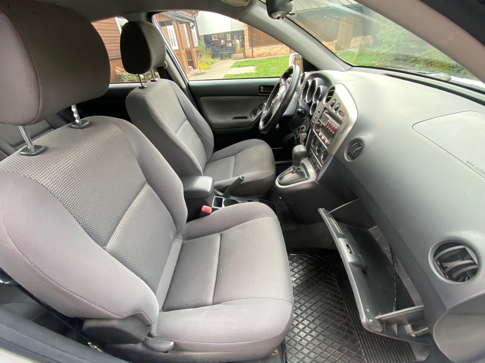 Wnętrze samochodu z szarą tapicerką, widoczne przednie fotele, deska rozdzielcza i otwarty schowek pasażera. Dywaniki gumowe z wzorem kratki. Widok z zewnątrz przez szybę.