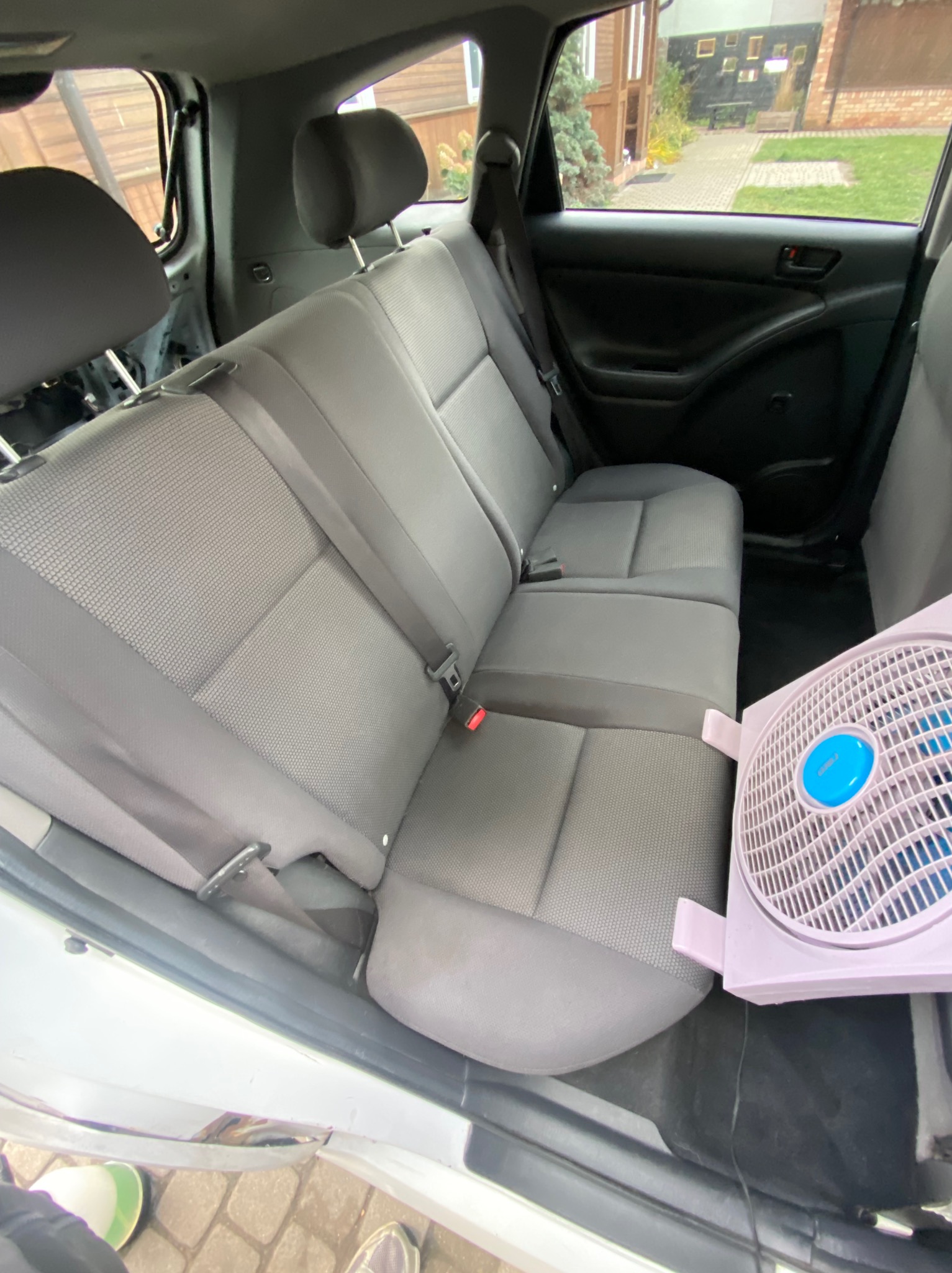 Wnętrze samochodu z szarą tapicerką, widoczne tylne siedzenia i fragment wentylatora, sugerujące proces suszenia po czyszczeniu.