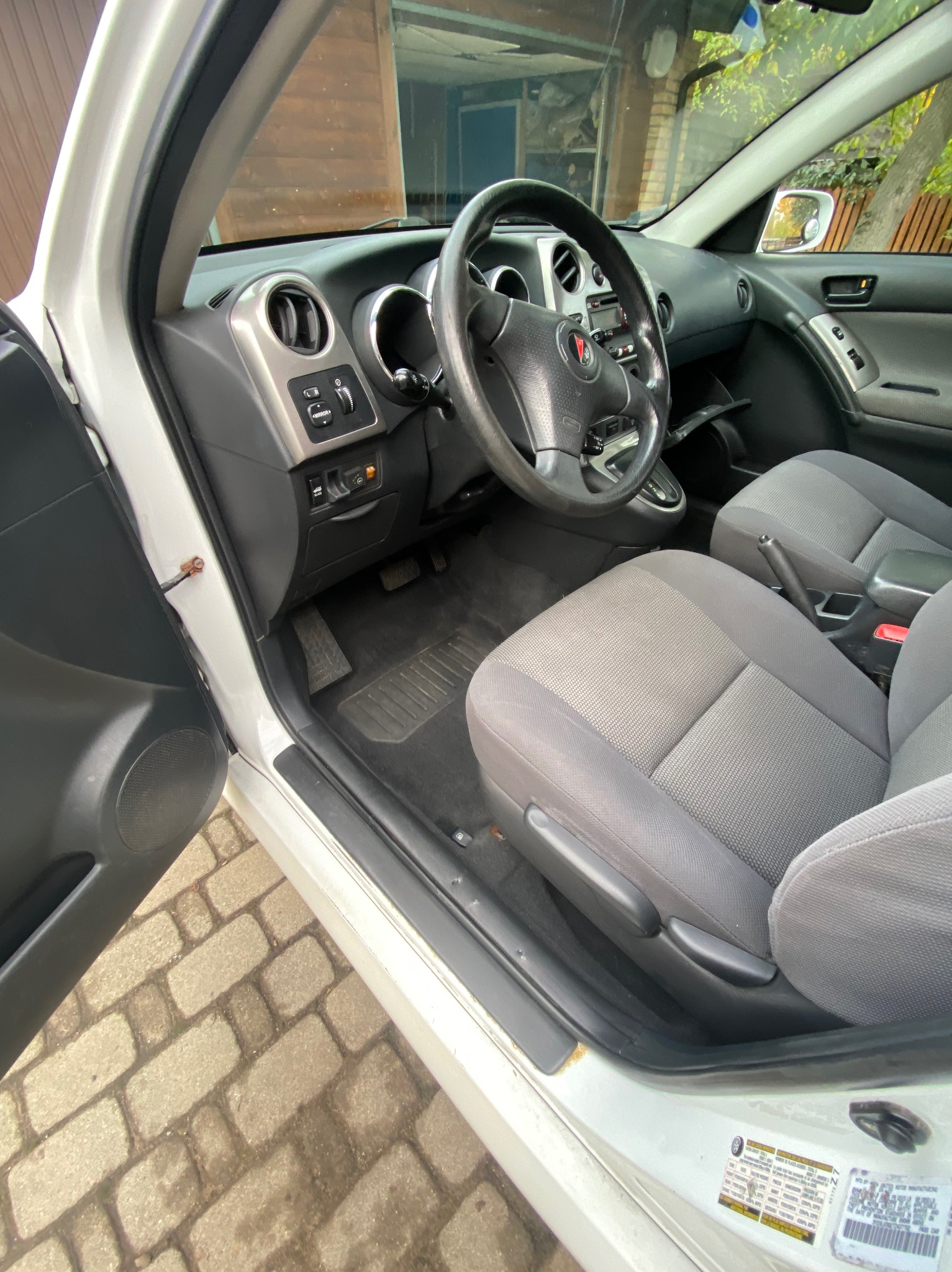 Wnętrze białego samochodu z otwartymi drzwiami kierowcy, widoczne szare fotele, deska rozdzielcza i fragment kostki brukowej na zewnątrz. Ujęcie z bliska, naturalne światło.