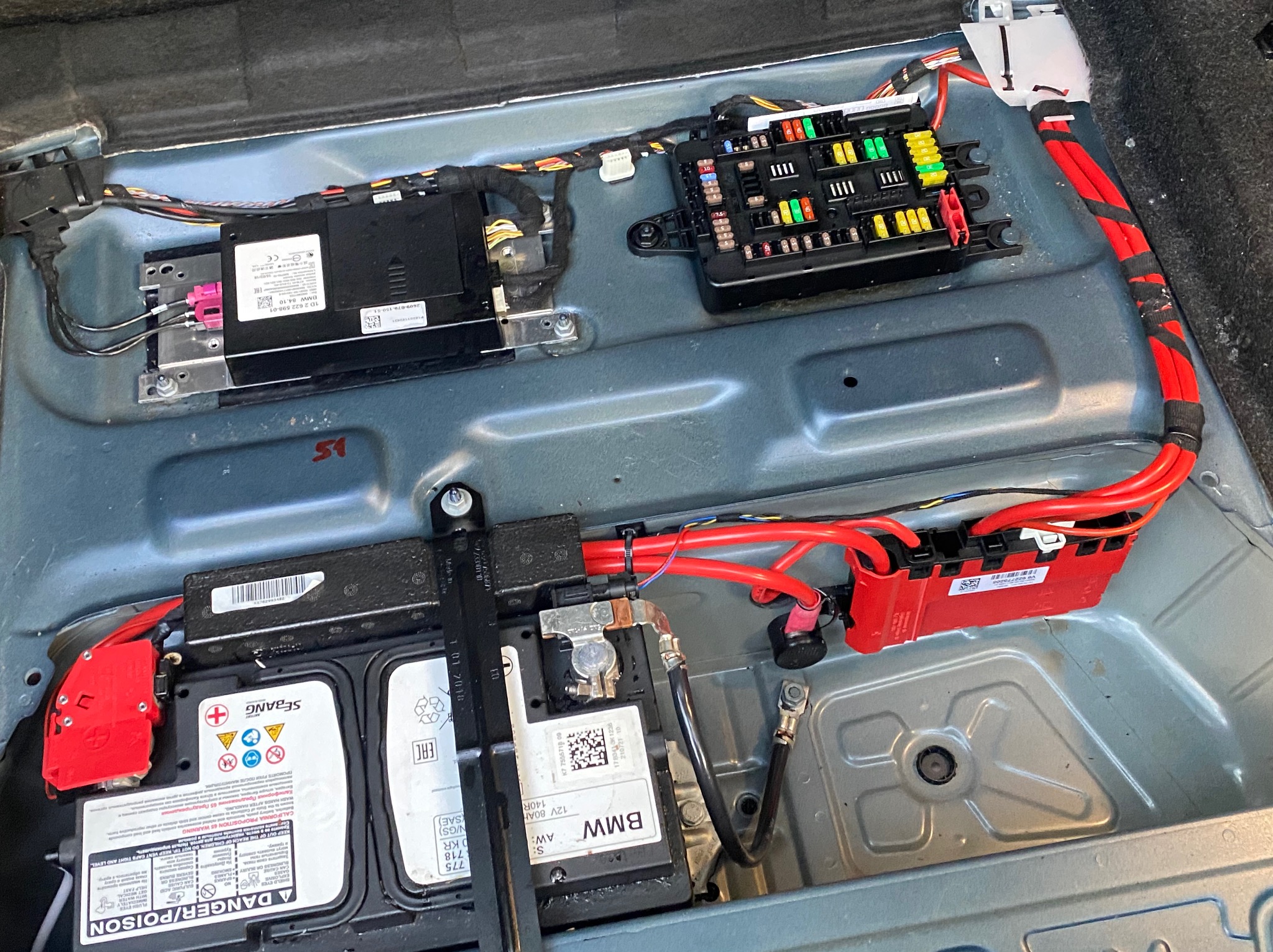 Wnętrze bagażnika BMW z widocznym akumulatorem, skrzynką bezpieczników i modułami elektronicznymi. Skupienie na detalach instalacji elektrycznej i komponentach.