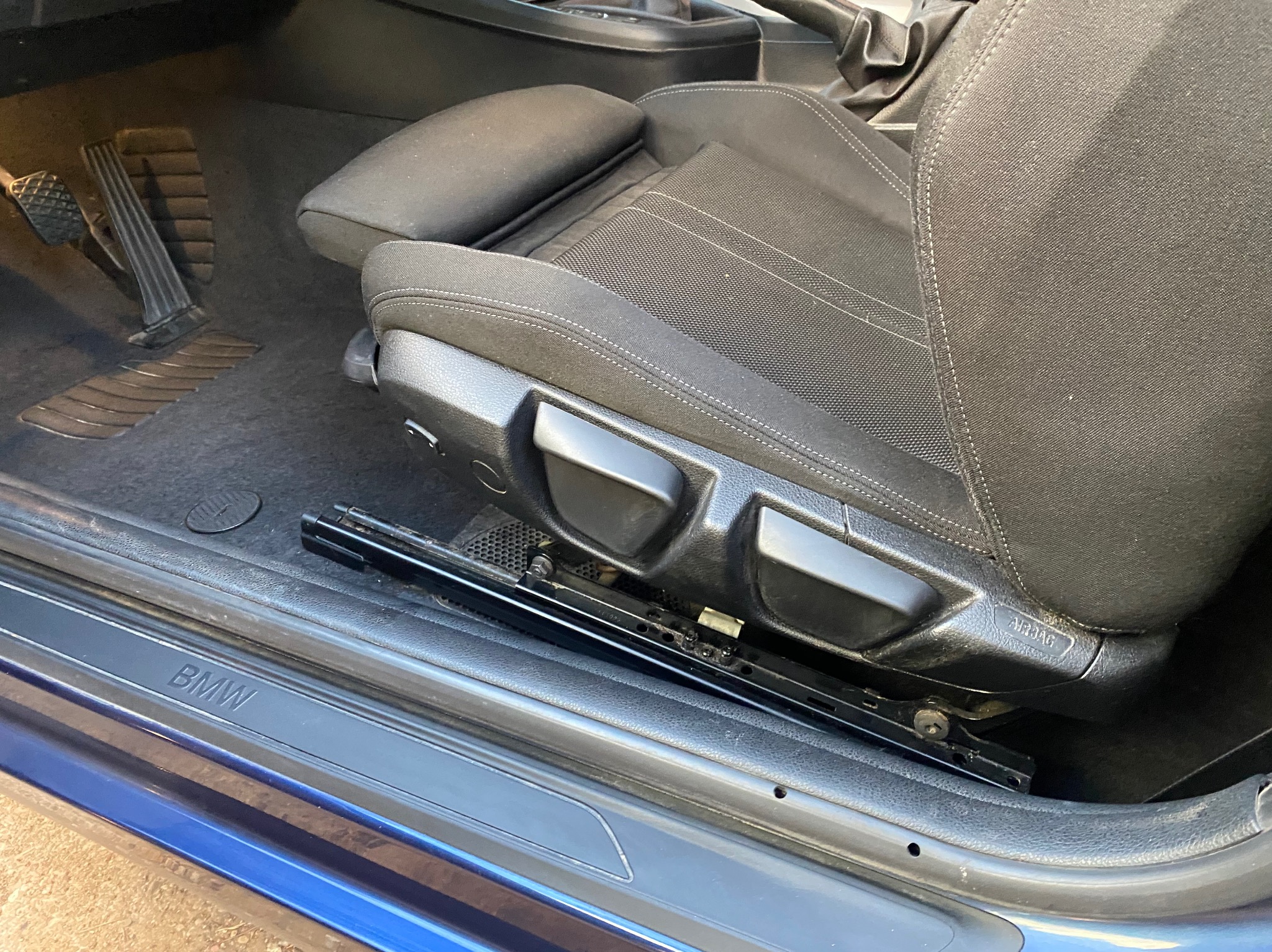 Czarne, tekstylne siedzenie samochodowe BMW z widocznymi pedałami i progiem. Detal wnętrza auta z bliska, ukazujący fakturę tapicerki i elementy regulacji fotela.