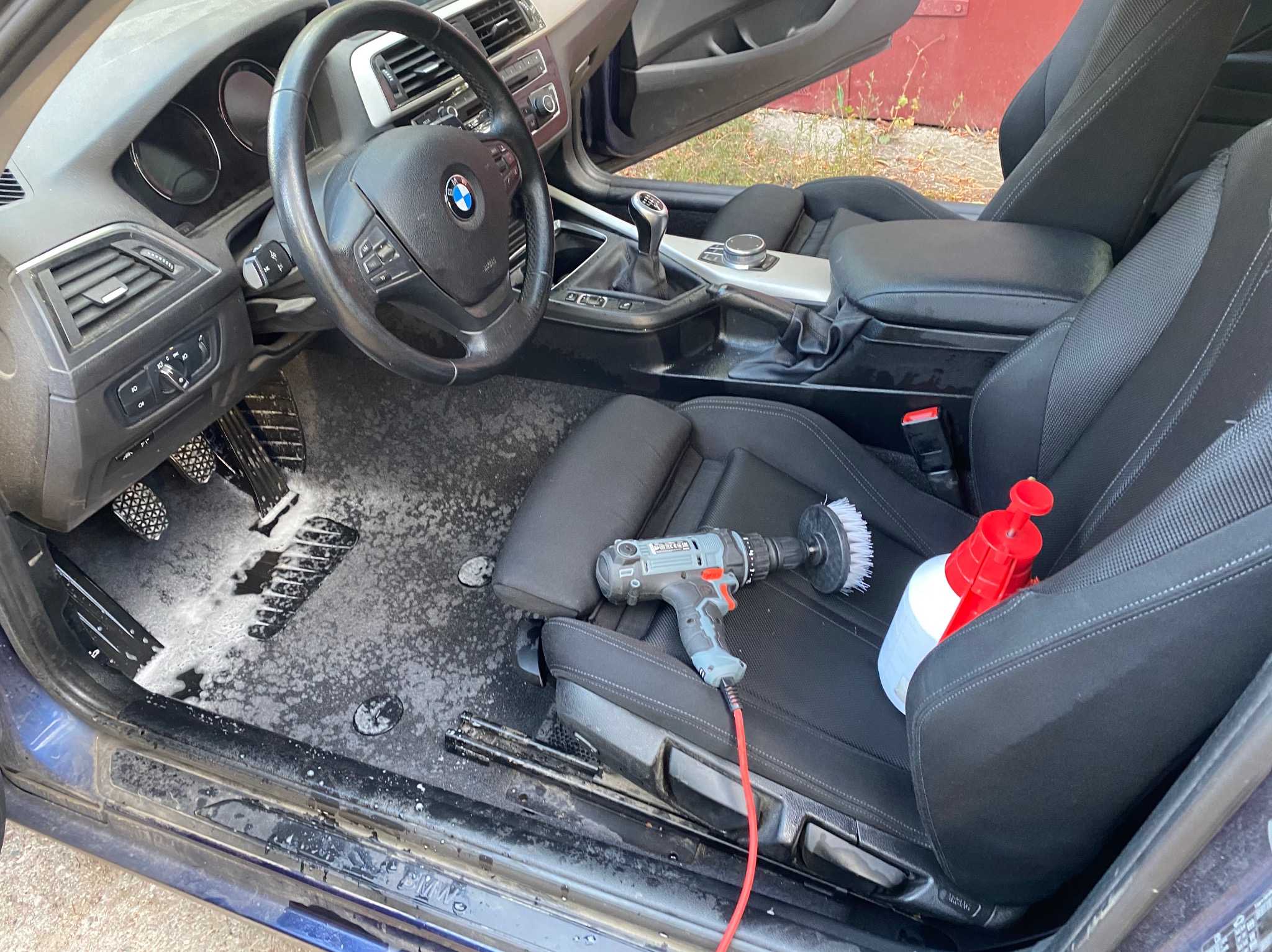 Wnętrze auta BMW w trakcie czyszczenia: piana na dywanikach, wiertarka z szczotką na siedzeniu, spryskiwacz. Widok z otwartymi drzwiami na deskę rozdzielczą.