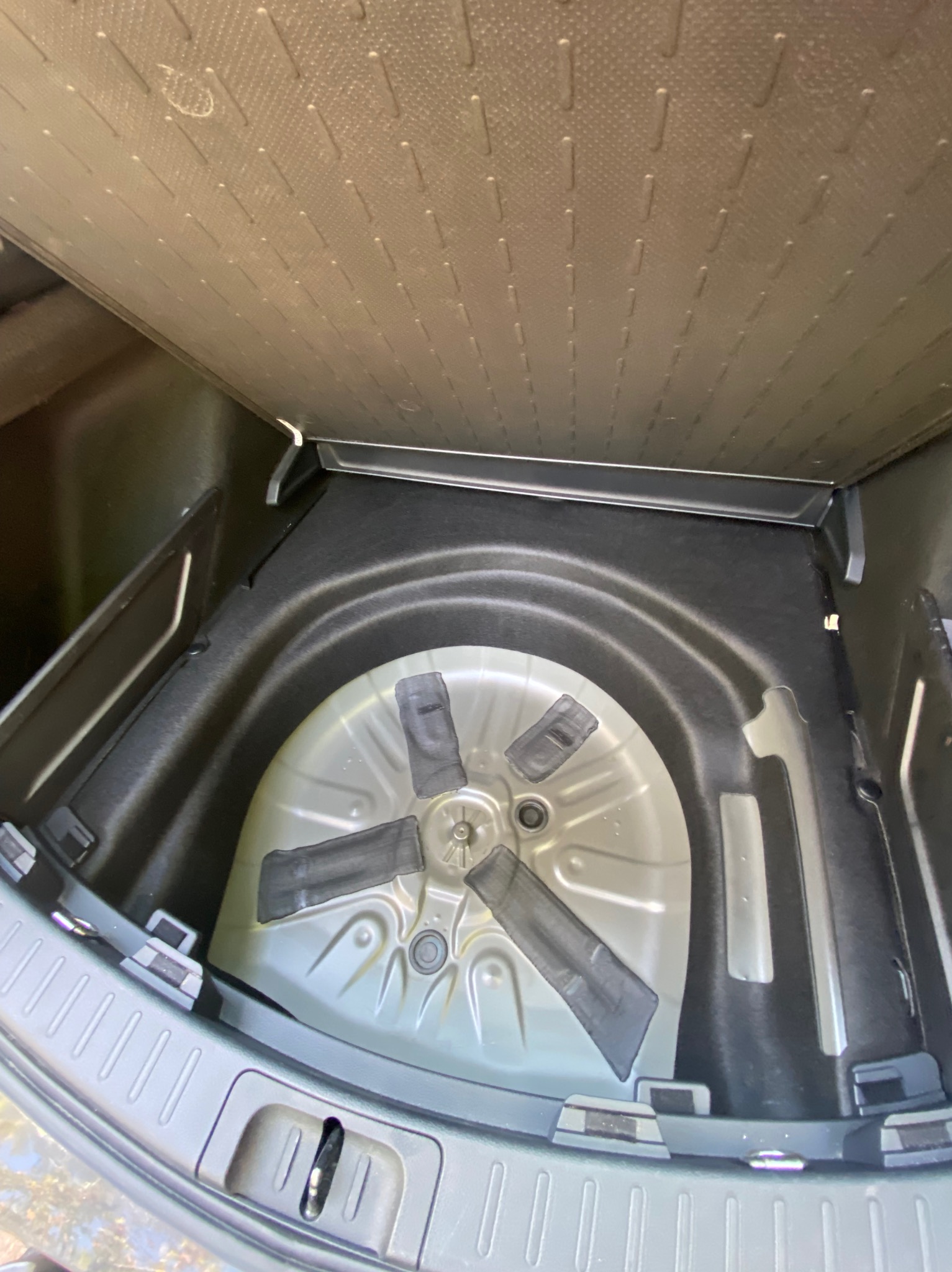 Wnętrze bagażnika samochodowego z podniesioną podłogą, ukazujące koło zapasowe i elementy mocujące. Widoczne detale wykończenia i faktura materiałów.