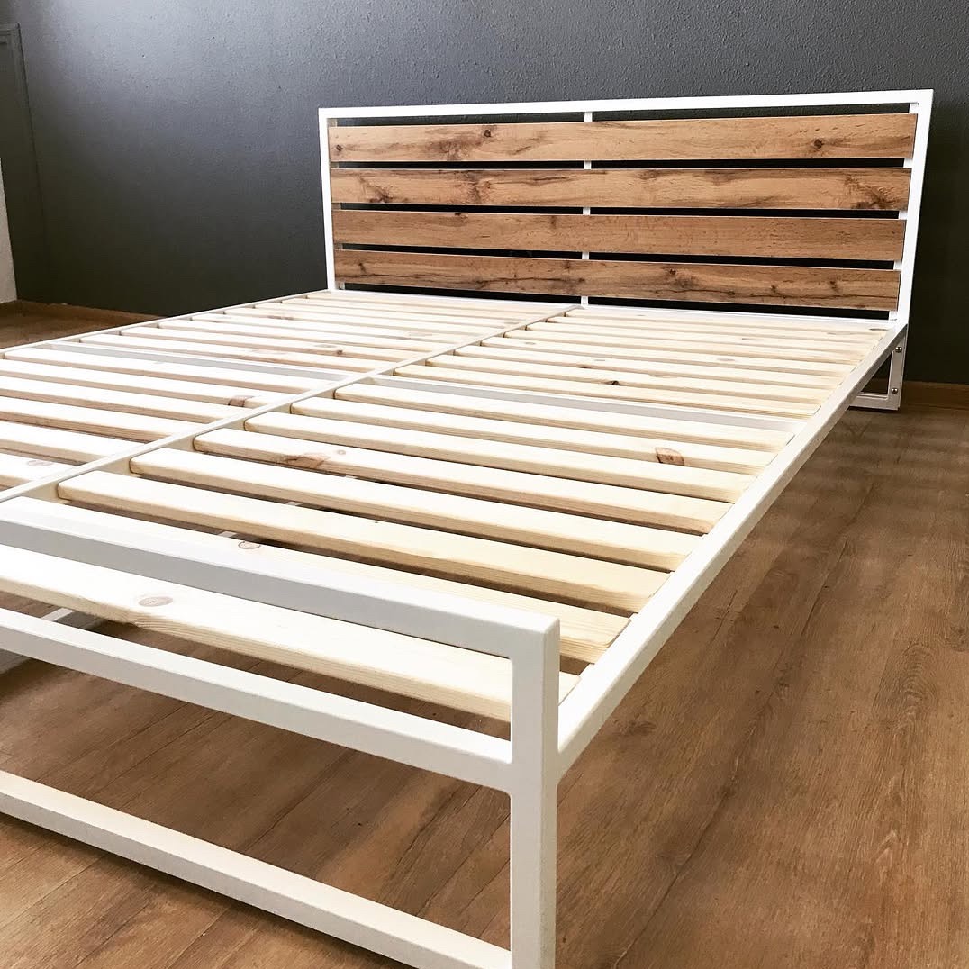 Metalowe łóżko z drewnianym zagłówkiem w stylu rustykalnym. Biała rama, drewniane listwy stelaża i zagłówek z desek o ciepłym odcieniu brązu. Minimalistyczny design.