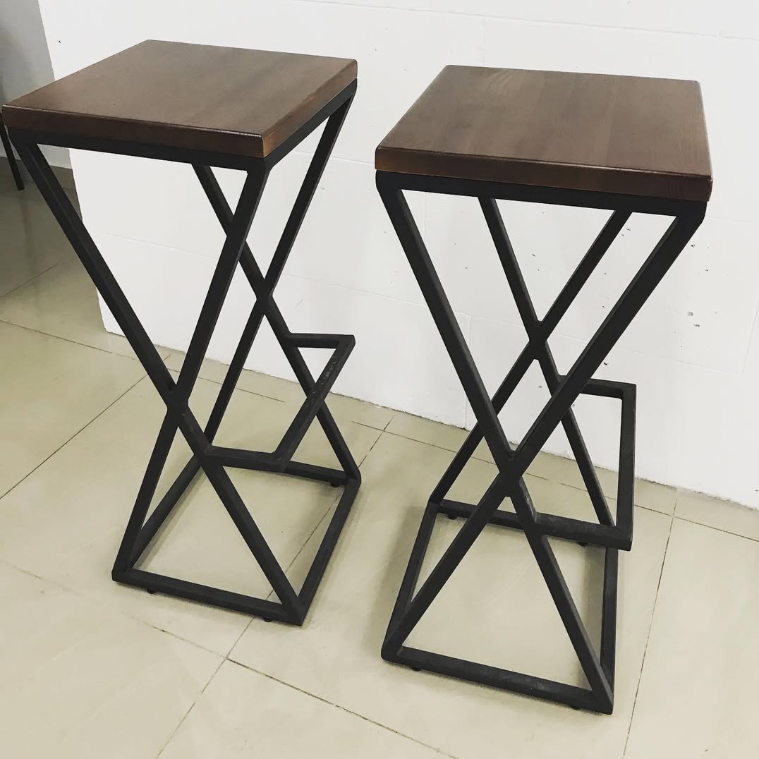 Dwa nowoczesne hokery z drewnianymi siedziskami i metalowymi, geometrycznymi podstawami w kształcie litery X, stojące na jasnej podłodze.