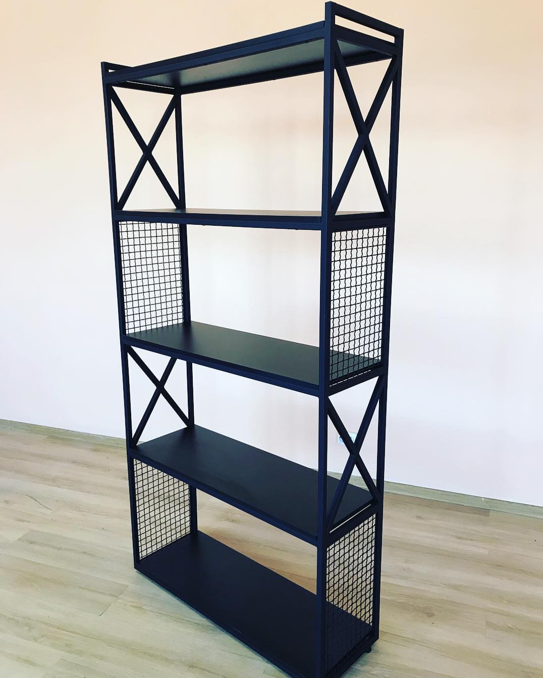 Czarny, metalowy regał z czterema półkami i siatkowymi bokami, ustawiony na jasnej podłodze, na tle kremowej ściany. Industrialny design.