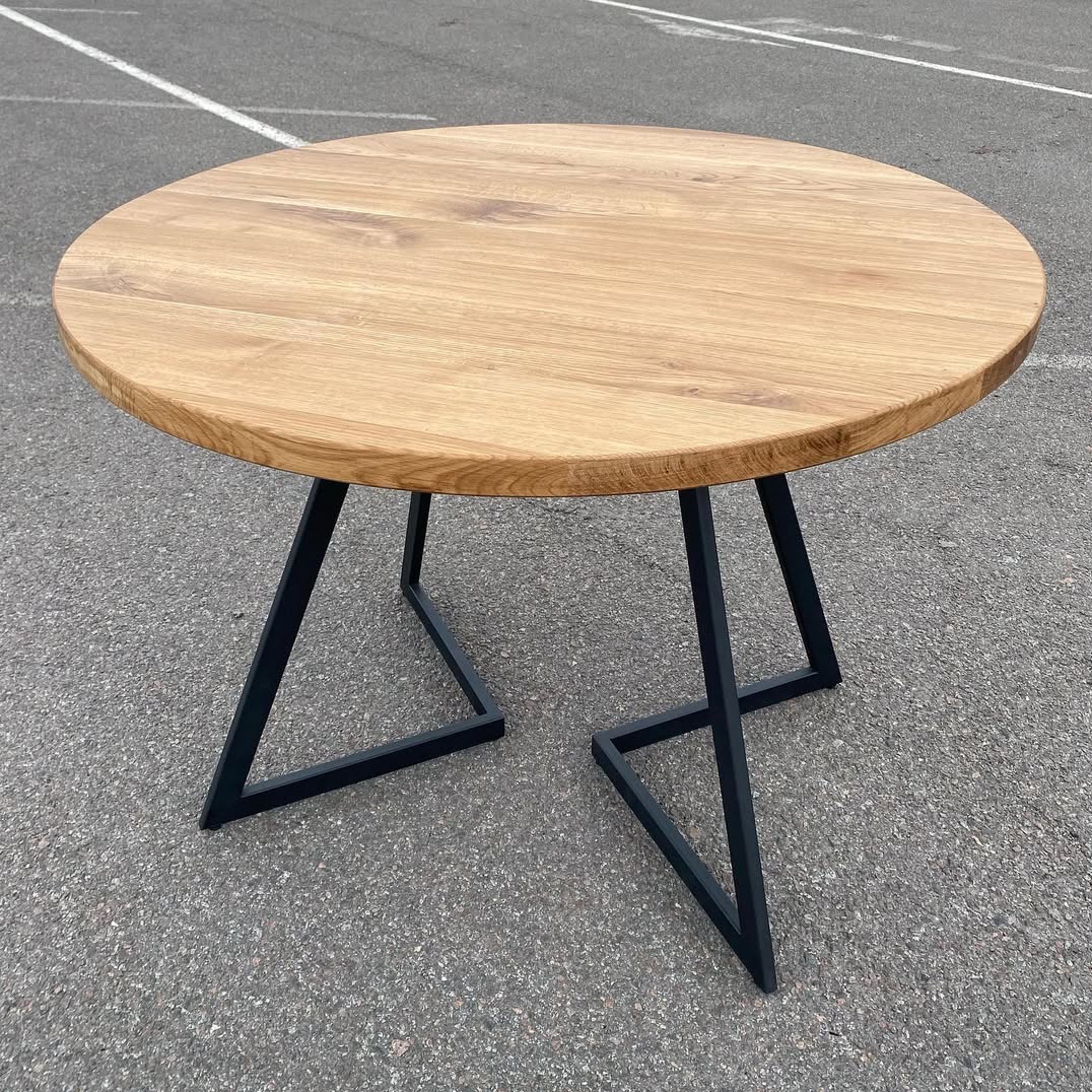 Okrągły stół z jasnego drewna na czarnych, metalowych nogach w kształcie litery V. Minimalistyczny design, widoczne usłojenie drewna, surowe tło.