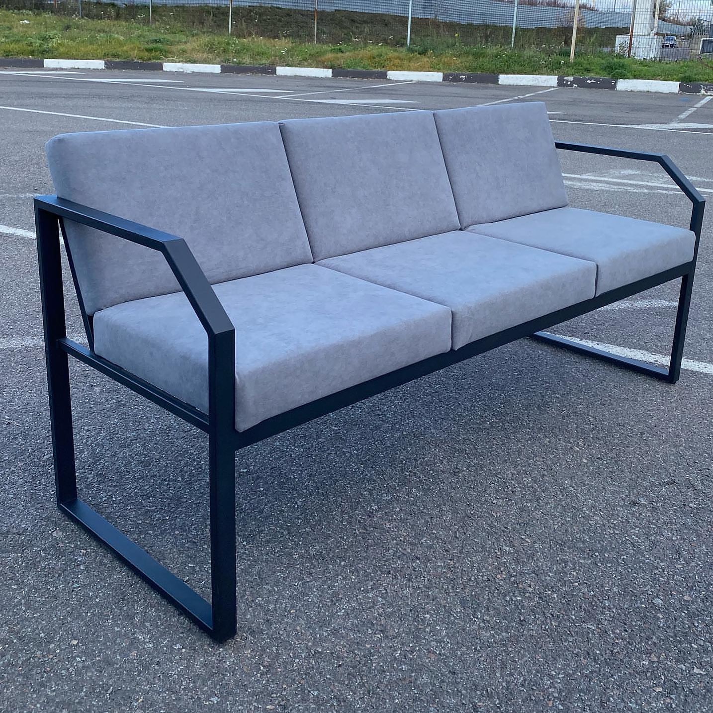 Metalowa sofa z szarymi poduchami na zewnątrz. Minimalistyczny design, spawana konstrukcja. Widoczne ślady spawania. Tło: parking z zielenią w oddali.