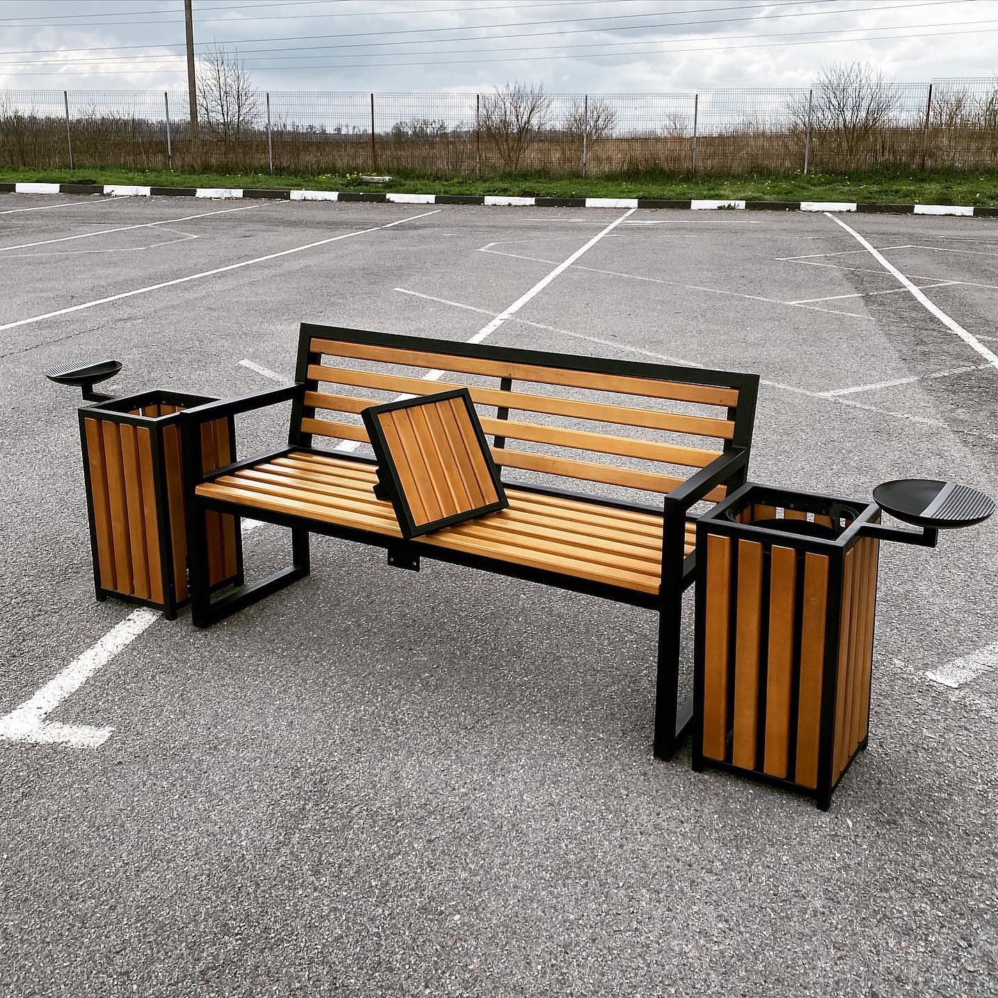 Metalowo-drewniana ławka parkowa z koszami na śmieci po bokach, na parkingu. Poduszka na siedzisku. Ujęcie z niskiej perspektywy, widoczne linie parkingowe.