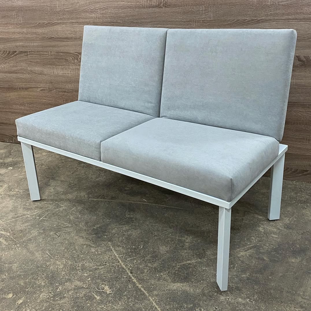Nowoczesna, dwuosobowa sofa z szarą tapicerką i białą, metalową ramą na tle drewnianej ściany i betonowej podłogi. Minimalistyczny design, proste linie.