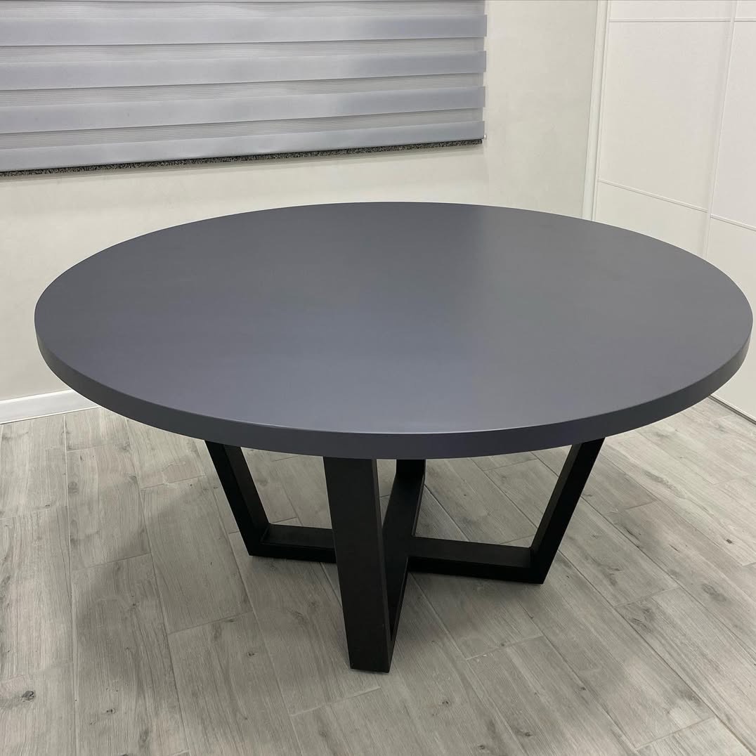 Szary okrągły stół na czarnej, geometrycznej podstawie w nowoczesnym wnętrzu z szarymi roletami i panelami podłogowymi. Minimalistyczny design.