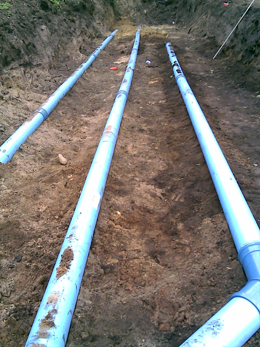 Trzy równoległe rury z tworzywa sztucznego, częściowo zakopane w wykopie ziemnym, widoczne z góry.