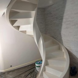 Usługi Remontowo - Budowlane Maciej Wiliński - Białe, spiralne schody po malowaniu, widoczne wiadro z farbą i zabezpieczona folią podłoga. Ściana wykończona dekoracyjną, białą cegłą.