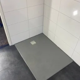 Nowoczesny brodzik z szarego kamienia, wkomponowany w białe płytki ścienne i ciemną podłogę. Minimalistyczny design, widoczne przyłącza wodne.