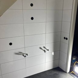 Usługi Remontowo - Budowlane Maciej Wiliński - Ściana wyłożona białymi, prostokątnymi płytkami z otworami na armaturę i gniazdka, oraz ciemna podłoga. Widoczne fragmenty instalacji wodnej i oświetlenie robocze.