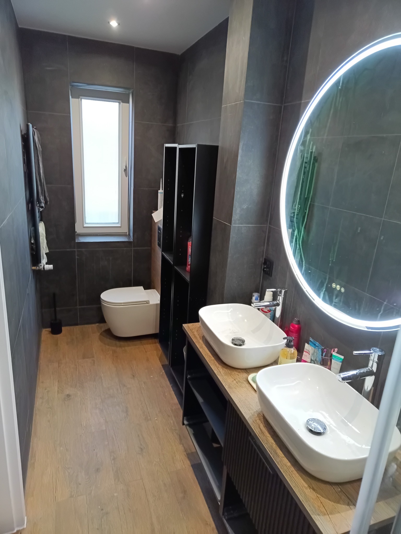 Nowoczesna łazienka z szarymi płytkami, drewnianą podłogą i podwójną umywalką z okrągłym lustrem LED. Widoczna toaleta i czarne półki.