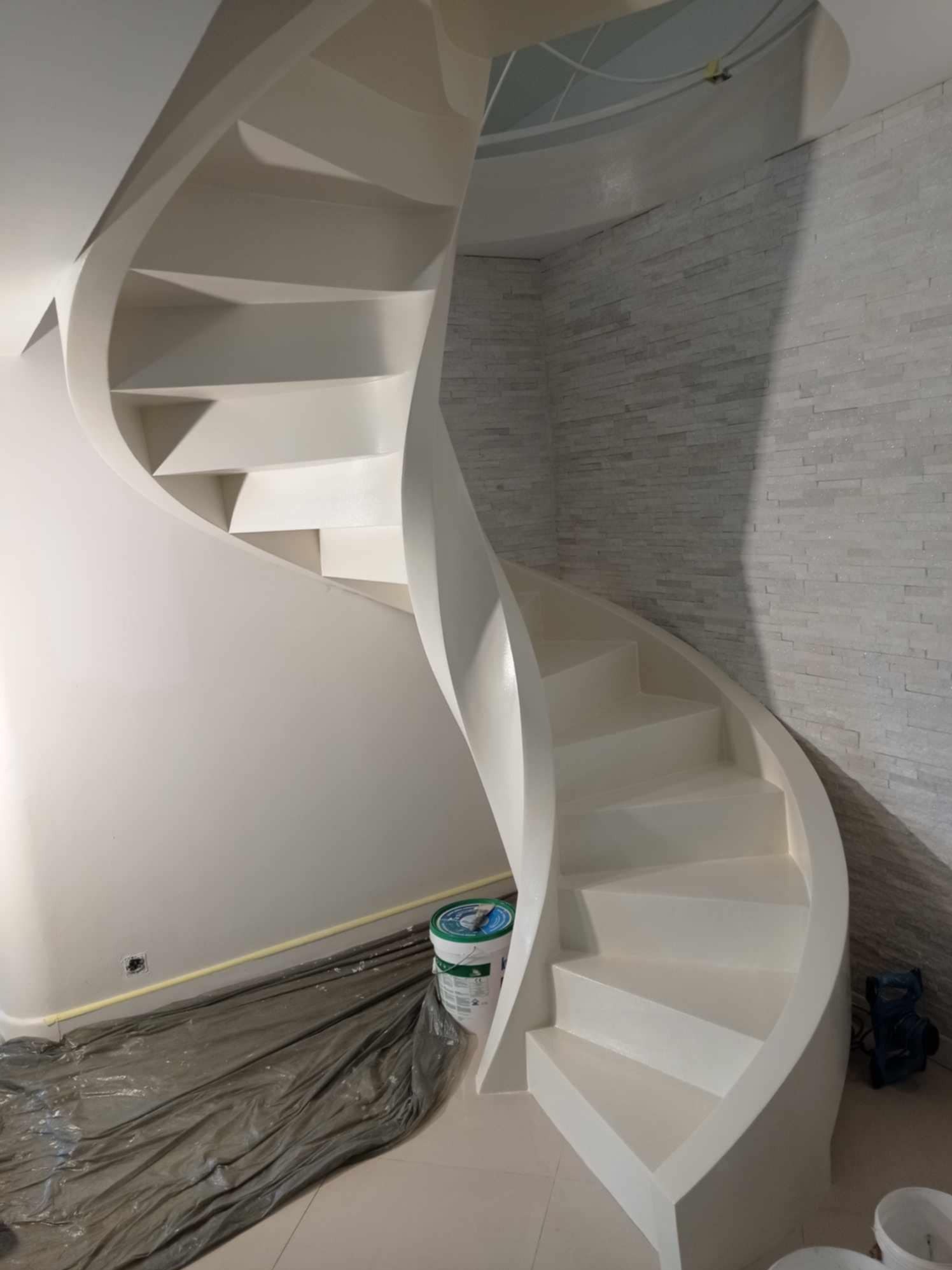 Białe, spiralne schody po malowaniu, widoczne wiadro z farbą i zabezpieczona folią podłoga. Ściana wykończona dekoracyjną, białą cegłą.