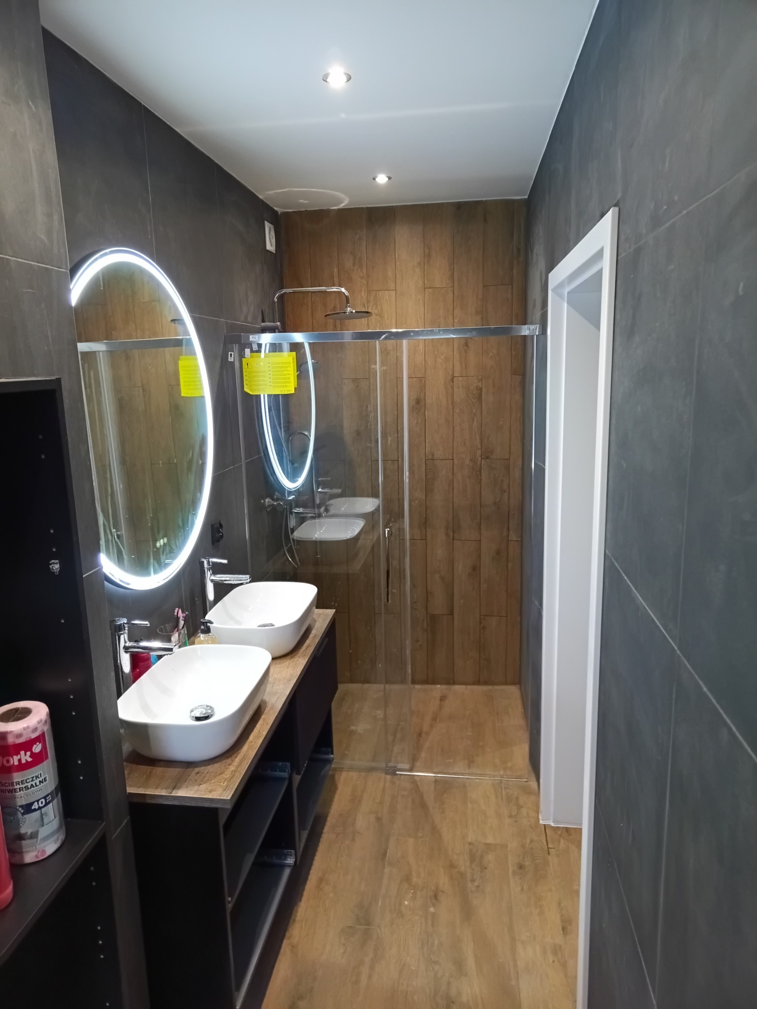 Nowoczesna łazienka z dwiema umywalkami nablatowymi, podświetlanym lustrem i kabiną prysznicową typu walk-in. Ciemne płytki kontrastują z drewnopodobną podłogą.