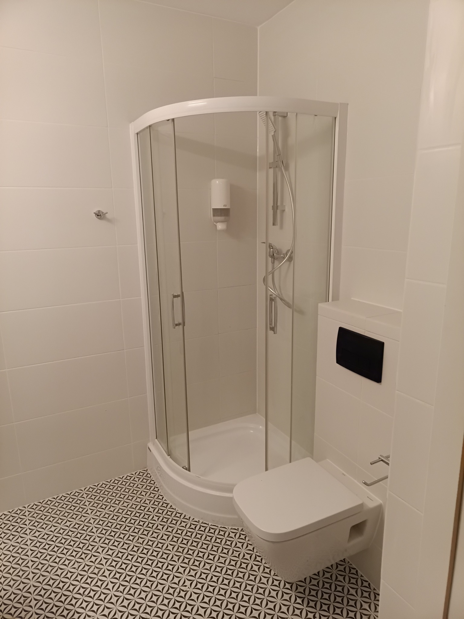 Biała łazienka z kabiną prysznicową i podwieszaną toaletą. Na podłodze wzorzyste, czarno-białe płytki. Minimalistyczny design, czyste linie, nowoczesne wykończenie.