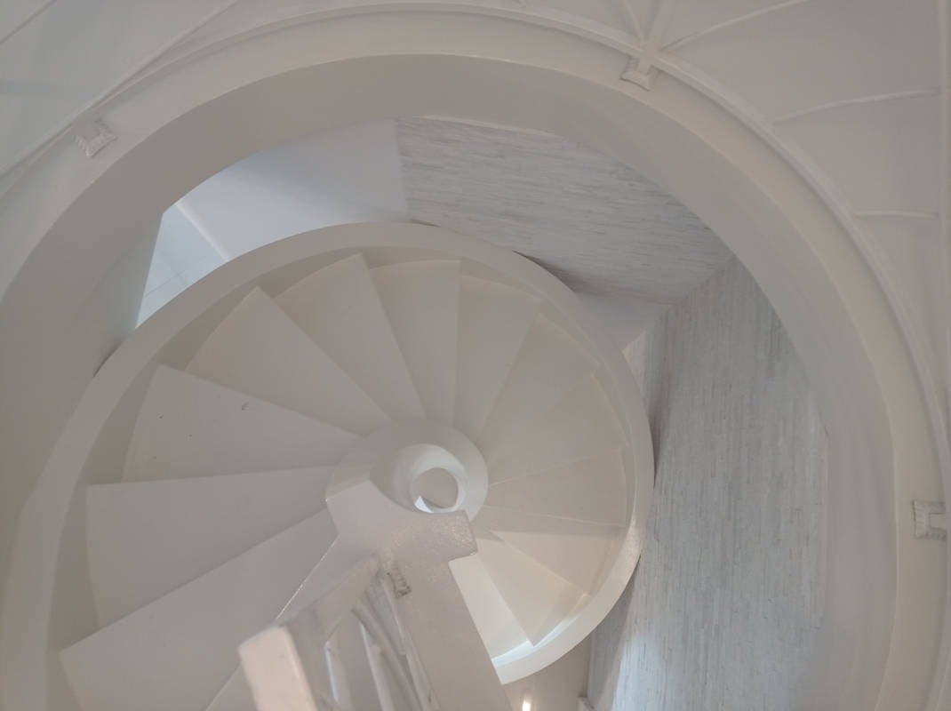 Białe, spiralne schody z góry, z widoczną strukturą ściany i sufitu. Minimalistyczny design, jasne oświetlenie, nowoczesne wykończenie wnętrza.