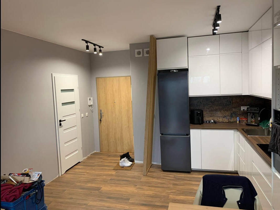Jasne wnętrze po remoncie: parkiet, białe drzwi z czarną klamką, nowoczesna kuchnia z białymi szafkami i czarną lodówką. Widoczne listwy przypodłogowe i oświetlenie.