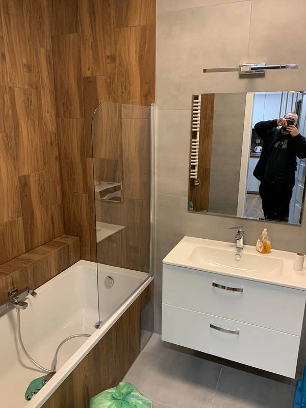 Nowoczesna łazienka z wanną i szklanym parawanem, drewnopodobne płytki na ścianie, biała szafka pod umywalką. W lustrze odbija się osoba robiąca zdjęcie.