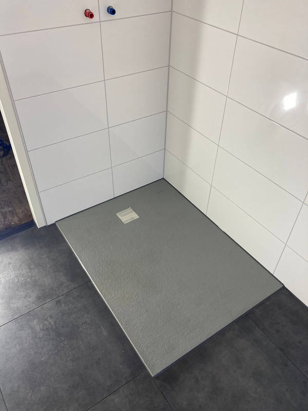 Nowoczesny brodzik z szarego kamienia, wkomponowany w białe płytki ścienne i ciemną podłogę. Minimalistyczny design, widoczne przyłącza wodne.