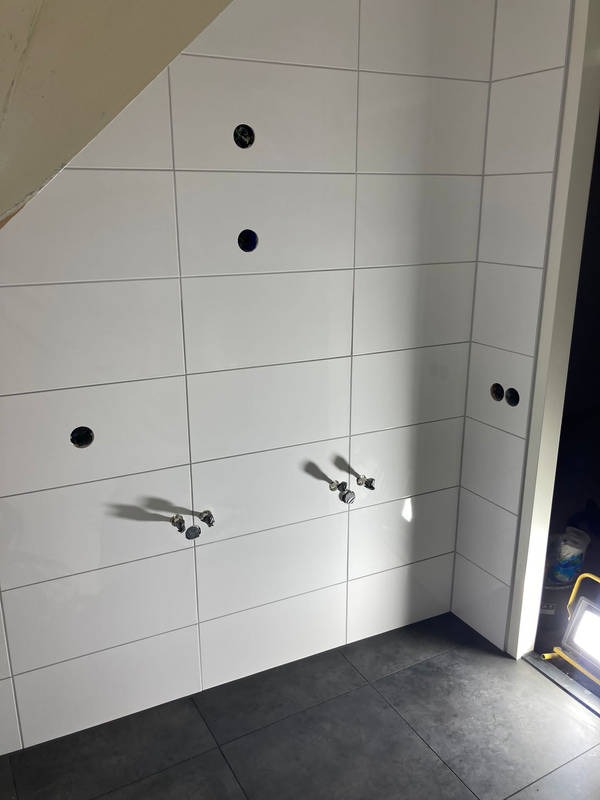 Ściana wyłożona białymi, prostokątnymi płytkami z otworami na armaturę i gniazdka, oraz ciemna podłoga. Widoczne fragmenty instalacji wodnej i oświetlenie robocze.