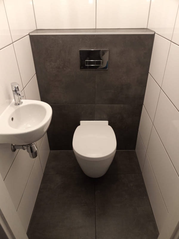 Kompaktowa łazienka z wiszącą toaletą i umywalką narożną. Szare płytki na podłodze i ścianie za toaletą, białe płytki na pozostałych ścianach. Chromowana armatura.